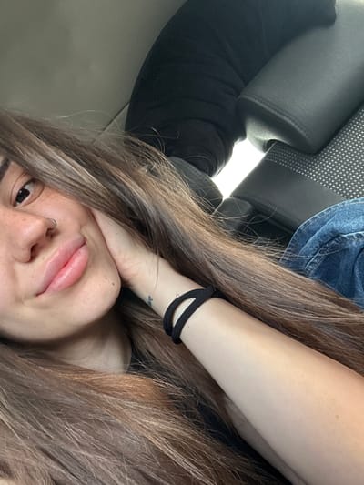 A close-up, rotated selfie shows a person with long, wavy brown hair, positioned on their left side with their head on the right of the frame, looking towards the camera. Their left arm is bent, with the hand resting near their chin. A black hair tie is worn on the wrist, and a faint, illegible tattoo is visible on the inner forearm. The person has a silver nose ring in their right nostril and slightly parted lips with a relaxed expression. One eye is clearly visible and open.
The setting appears to be inside a vehicle, suggested by the dark grey car seat headrest and a portion of a patterned car seat backrest in the background. A piece of blue denim fabric is also visible, likely part of clothing. The lighting indicates it is daytime. The overall mood is casual and relaxed, capturing a moment within a car in Aldomirovtsi, Bulgaria. No legible text is visible within the image beyond the unreadable tattoo.