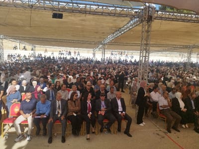 The image captures a large outdoor gathering in Al-Khalil, Palestinian Territory. Hundreds of individuals, primarily adults, are seated in rows of red and gold chairs under an extensive temporary canopy. The canopy consists of a light-colored fabric roof supported by a structure of metal trussing and vertical poles. The audience, a mix of men and women, many of whom wear hijabs, are oriented forward, consistent with attending an event. Attire varies from casual to semi-formal, with men in button-down shirts and suits, and women in modest clothing. Visible objects include a speaker suspended from the trussing. In the mid-ground, a man holds what appears to be a microphone or small camera. Further back, another individual is seen near a camera on a tripod. Beyond the immediate seating area, additional people are visible standing on a sloped, unpaved terrain. The scene is illuminated by bright daylight, indicating an outdoor setting.