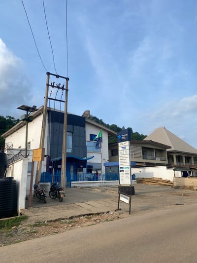 This detailed image captures a street scene in Igarra, Nigeria, under a bright, partly cloudy sky. The primary focus is a two-story building identified as "Keystone Bank," featuring a blue and white facade with a prominent vertical sign. The bank sign lists "ATM 24/7" and various services including "MoneyGram," "Wari," "DCS," and "Remitly," alongside different account types. A Nigerian flag (green and white) is mounted on the bank's exterior.

To the left of the bank, a white building with a gray roof and barbed wire atop its perimeter wall is visible. A tall pole in front of this building supports a solar panel and associated equipment. A large black water tank sits on the ground near the left edge of the frame. In front of the bank, a blue metal fence borders a paved area where two motorcycles are parked. A "MoneyGram" sign is affixed to the side of the bank building.

Dominating the central foreground is a substantial wooden utility pole with multiple electrical lines, insulators, and what appears to be a transformer or junction box. These lines extend upwards towards the sky.

To the right of the bank, an older two-story building with a multi-textured facade and a pyramid-shaped roof is visible. Further right, a pile of bamboo or wooden poles lies on the ground next to a small corrugated metal structure. The foreground features an asphalt road with a "NO PARKING" sign on a stand near the curb. The ground surrounding the road is uneven, with visible dirt, gravel, and sparse vegetation.

In the background, lush green hills rise, crowned by distinctive rocky outcrops. One person wearing a red shirt is partially visible near the bank's entrance on the right side of the blue fence.
