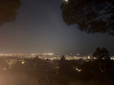 The image captures a nighttime panoramic view of a coastal city, likely from an elevated vantage point. In the foreground, dark silhouettes of trees and foliage obscure the lower portion of the scene. Beyond the trees, the city lights stretch across the landscape, indicating a densely populated area. Numerous buildings are visible, illuminated by their internal lights. Further in the distance, the lights suggest the presence of a harbor or port, with several vessels illuminated on the water. A prominent structure with a distinctive illuminated spire stands out amongst the city lights. The sky above is dark, with a subtle haze or fog present, and a bright circular object, possibly the moon, is visible in the upper right quadrant. The overall atmosphere is nocturnal and somewhat atmospheric due to the haze and low light conditions. The location is identified as the city of Agios Pavlos Municipal Unit, Greece. There are no people visible in the image.
