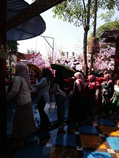 The image depicts a street scene in Tegalwangi, Indonesia, characterized by a bright, sunny sky and lush greenery. In the foreground, several individuals are observed, predominantly women wearing headscarves and modest attire. One woman in the left foreground wears a light-colored, vertically striped long-sleeved shirt and a long, light brown skirt. To her right, another woman is visible from the waist down, wearing a dark, flowing garment and dark pants. Further to the right, a woman in a light blue, long-sleeved shirt and dark pants is seen with her back to the camera.
Behind the people, a vibrant display of artificial cherry blossoms in various shades of pink adds a festive atmosphere. Red railings line the area, separating the onlookers from a colorful tiled floor, which appears wet and reflects the sunlight. Several small, round objects, possibly toys or balls, are scattered on the tiled surface.
In the background, traditional Indonesian architecture is evident, with red buildings featuring ornate details and curved roofs. A tall tree with branches laden with green leaves stands prominently, and a rustic, cylindrical woven lantern hangs from one of its branches. The overall scene suggests a public gathering or a cultural event, possibly a temple or a recreational area, given the decorative elements and the presence of children in the middle ground. The atmosphere is lively and engaging, with a blend of cultural and natural elements.