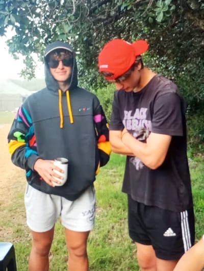 The image displays two male individuals standing outdoors against a backdrop of green foliage. The subject on the left wears a dark grey hooded sweatshirt with multicolored sleeves (teal, purple, black, yellow, orange), light-colored shorts with a logo, sunglasses, and a cap. This individual holds a white thermal mug in the right hand and looks forward with a slight smile. The subject on the right wears a backward red baseball cap, a dark t-shirt bearing "BLACK SABBATH" text, and black Adidas athletic shorts with white side stripes. This individual stands with arms crossed, gazing downwards. The ground is composed of dirt and sparse grass. A dark, low object is partially visible in the bottom left. A faint, distant structure is visible in the background behind the left subject. The image provides no visual details to confirm the location as Aktash, Uzbekistan.