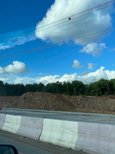 The image shows a view of the sky, clouds, trees, and what appears to be a road construction site in Novodrozhzhino, Russia. Power lines with a red marker are visible against the blue sky. White clouds dot the sky. A row of trees is adjacent to a mound of dirt. There is a gray concrete barrier along the road. The image seems to be taken from within a vehicle.