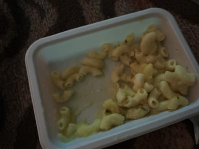 A white, rectangular plastic container holds a portion of macaroni and cheese. The macaroni pieces are curved and appear to be coated in a creamy, yellowish cheese sauce. Some pieces of dark green herb are visible mixed within the pasta. The container is resting on a dark, textured surface, possibly a blanket or rug, which has a woven pattern. The lighting in the scene is dim, casting soft shadows and highlighting the sheen of the sauce on the pasta. The image is cropped such that only the food and its immediate surroundings are visible. There is no indication of location within Merrylands, Australia, or any people or actions.