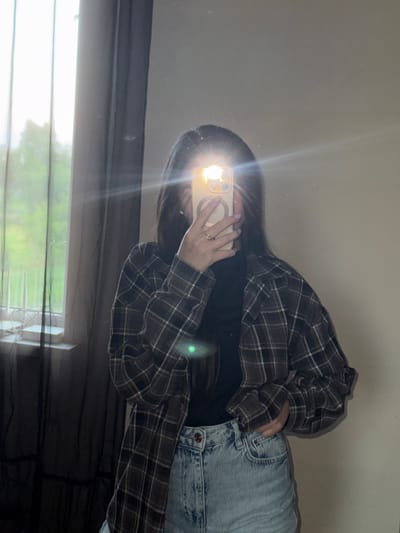 An overhead shot captures a young woman standing in front of a mirror, taking a selfie in what appears to be her bedroom. She is wearing a plaid jacket over a black turtleneck and denim shorts. The room has a window with dark curtains and a view of greenery outside, suggesting it is located in Horenychi, Ukraine. The lighting is dim, with a bright glare from the phone's flash obscuring the woman's face.