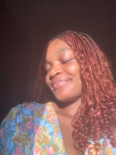 A close-up, chest-up portrait features an individual identified as female, with dark skin, facing slightly left of center, eyes closed, and a slight smile. Her hair is styled in numerous small, red-brown box braids, some ending in curly strands, falling over her shoulders. She wears a garment with a floral pattern in shades of light blue, yellow, and pink. Strong, directional lighting illuminates the right side of her face and shoulder, casting bright highlights. The background is uniformly dark and indistinct. The scene is located in the city of Anantigha, Nigeria.
