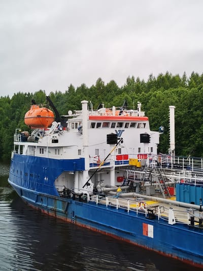A blue and white tanker ship is docked in the city of Volokov Most, Russia, with lush green trees lining the background. The ship has an orange life raft suspended from a crane on its upper deck. Various pipes, ladders, and safety equipment are visible on the ship's deck. The water surrounding the ship is dark and reflects the overcast sky. The overall scene depicts a serene, industrial setting within a natural landscape.