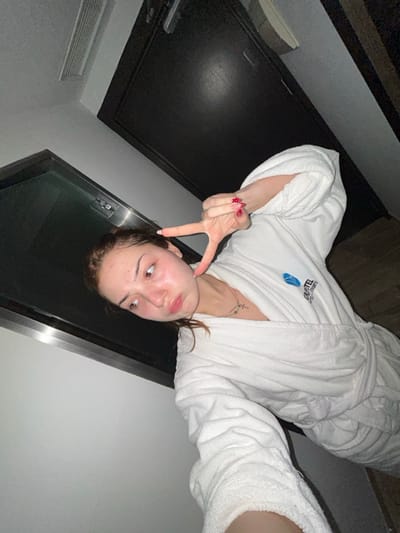 A woman in a white bathrobe poses for a photo, upside down relative to the viewer. She gives a peace sign with her left hand, displaying red patterned nails. She wears a necklace with a cross. The background features a dark door, a wall with an air vent, and a large window. The bathrobe has writing on the left breast. Her skin appears fair and her hair is pulled back.