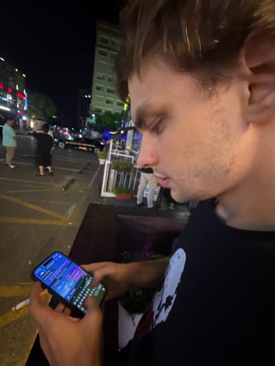 The image depicts a nighttime urban scene, likely in Minhang District, China, characterized by artificial lighting. In the foreground, a person is partially visible, oriented sideways. This individual has short brown hair and light stubble, and is wearing a dark t-shirt, looking downwards.

Adjacent to them, another person's hands are prominently featured, actively engaged with a smartphone. The phone screen is brightly illuminated, displaying a messaging application interface with blue/purple chat bubbles and a virtual keyboard where the user's thumbs are positioned as if typing. A lit cigarette is held between the fingers of the hand operating the phone. This individual is wearing a black t-shirt adorned with a distinctive white graphic on the chest, possibly an abstract design or a stylized figure.

The background shows a busy city street after dark. Multi-story buildings with numerous lit windows line the street, indicating a vibrant urban environment. Several blurry figures of pedestrians are seen walking or standing, suggesting ongoing activity. A black car is visible, and the paved ground features yellow road markings. The overall setting portrays an active evening in a metropolitan area. The image itself is rotated 90 degrees clockwise. Due to the angle and resolution, specific text on the phone screen is not clearly discernible.