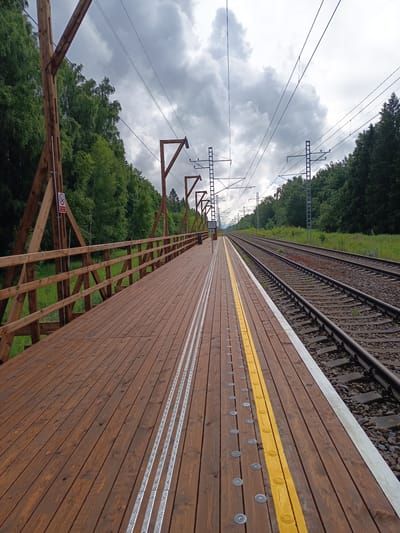 The image depicts a wooden train platform at a rural station, likely in Kulli, Estonia, under a cloudy sky. The platform is made of weathered wooden planks, with a yellow safety line marking the edge nearest the tracks. Raised tactile warning strips, appearing to be aluminum, run parallel to the yellow line. Circular metal discs are embedded at intervals along the platform surface.
To the right of the platform, a set of train tracks runs parallel to the platform, curving slightly into the distance. The tracks are laid on ballast, with railway signals and overhead power lines visible in the background.
The left side of the platform is bordered by a wooden fence and supported by wooden posts and diagonal beams, characteristic of a rustic or perhaps newly constructed platform. Beyond the fence, dense green trees and foliage form a natural backdrop, suggesting the station is situated in a wooded area. The sky is overcast with dramatic, dark clouds. There are no people or animals visible in the scene. The overall impression is one of a quiet, somewhat isolated rural train station.