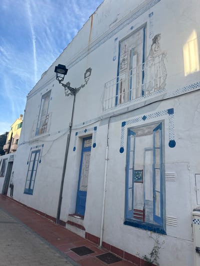 The image displays a white building facade in Calella De Palafrugell, Spain, extensively adorned with blue and white trompe l'oeil murals under a clear blue sky with faint contrails. A prominent black, ornate street lamp with two light fixtures is affixed to the upper half of the wall, left of center.
On the upper level, painted architectural features include two windows on the left and a central balcony with an open door, through which a female figure in a white dress leans on the railing, observing a small white bird on the balcony floor. Blue checkered and circular patterns border these painted elements and form horizontal decorative lines across the facade. Faint rust streaks are visible on the upper wall.
The lower level features three distinct painted sections: two blue windows on the far left, a central blue door with white ornamental detailing, and a large painted open window on the right revealing an interior scene with a red chair and a framed picture. Vertical white pipes run along the wall between these elements. The ground level presents a reddish-brown tiled sidewalk with two dark rectangular doormats. A small plant with yellow flowers grows at the base of the wall on the right. A partial view of another light-colored building is visible in the background on the far left.