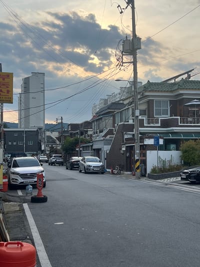 The image shows a street scene in a city, likely Gwangju, South Korea, with a mix of residential and commercial buildings. The sky is overcast with patches of sunlight breaking through the clouds, and there are numerous overhead electrical wires crisscrossing the scene. On the left side of the street, a white SUV is parked, and a red traffic cone is placed in front of it. Further down the street, several other cars are parked and driving. The buildings vary in architectural style, with some appearing to be older, brick structures with tiled roofs, and others being more modern, multi-story buildings. There are also streetlights and utility poles visible. The street itself is paved, and the overall impression is of a typical urban neighborhood. A person can be seen walking on the sidewalk in the distance.
