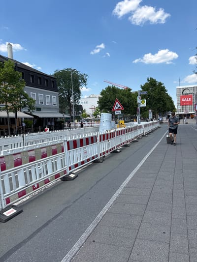 The image shows a street scene in Erlangen, Germany, during daytime under a partly cloudy sky. Construction barriers with red and white reflective stripes line the left side of the street, separating the road from a sidewalk. A person is walking on the sidewalk on the right, pulling a dark grey suitcase. Behind the construction barriers, there are buildings, trees, and street signs. One sign indicates "Rathausplatz". A red construction crane is visible in the background. Another building in the background has a large "GALERIA SALE" sign. The street appears to be undergoing some form of roadwork or renovation.