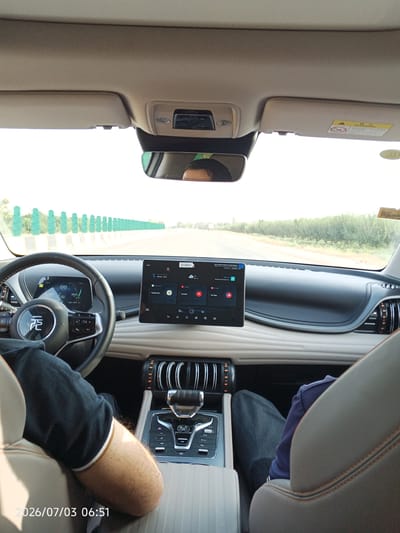 The image shows the interior of a car, viewed from the perspective of the passenger seat. The dashboard features a large touchscreen display and a digital instrument cluster. The steering wheel is visible, with the BYD logo prominently displayed. The gear selector and other car controls are also visible. The car appears to be in motion, as indicated by the blurred scenery visible through the windshield. The scenery consists of a road flanked by green vegetation and a sky with white clouds. The time stamp in the bottom left corner indicates July 3, 2026, at 06:51. Based on the metadata, this image was taken in Kiikmazor, Tajikistan.