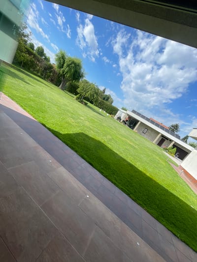 The image depicts an outdoor scene with a house, lawn, and patio. The lawn is green and well-maintained, with trees bordering the left side of the frame. A house is visible in the background with solar panels installed on the roof. To the right of the lawn is a tiled patio area. The sky is blue with scattered clouds. The image appears to be taken from indoors, looking out through a window or doorway. The location is Ryahovo, Bulgaria.