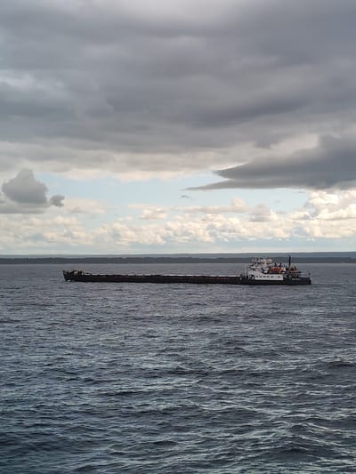 A barge is traveling on the water near Ozernoe Ust'e, Russia. The sky is overcast with dark clouds. The water is choppy and dark blue. The barge is long and dark, with a white superstructure at the stern. The superstructure has windows and railings. There are several life rafts on the deck of the barge. The horizon is visible in the background, with a line of trees. The water takes up most of the foreground of the image.
