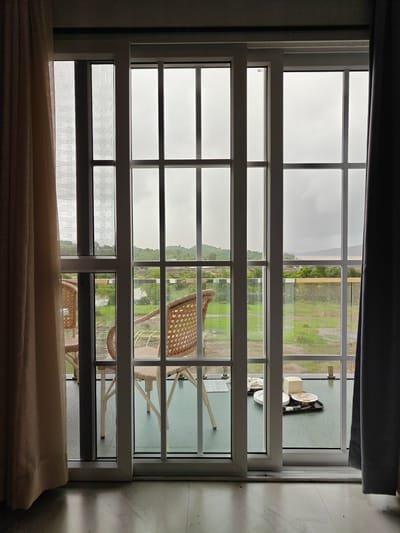 A view through a sliding glass door reveals a balcony with two wicker chairs. On the balcony, a tray holds two plates with food, one plate with two fried eggs and bacon, and another plate with a stack of three white plates, a small white box, and silverware. In the background, a body of water is visible, surrounded by lush green hills under a cloudy sky. The scene is located in Ajivali, India.
