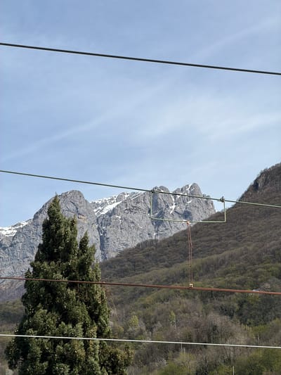 The image shows a mountain landscape with power lines running across the foreground. The location is Mandello del Lario, Italy. A gray, rocky mountain with patches of snow is visible, partially obscured by a green tree in the lower right corner. A forested hillside borders the mountain on the right side. The sky is light blue with wispy clouds. A green and a reddish-brown power line run vertically across the frame. The angle of the photo is skewed.