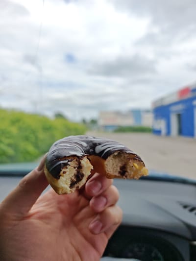 A hand is holding a half-eaten chocolate-frosted donut with white icing drizzle. The donut has a bite taken out of it, revealing a light-colored dough with dark chocolate filling. The background is blurred, showing a cloudy sky, green foliage, and a blue building with white trim, suggesting an outdoor urban or semi-urban setting. The scene appears to be captured from inside a vehicle, as indicated by the dashboard visible in the foreground. The location is identified as Cheremkhovo, Russia.