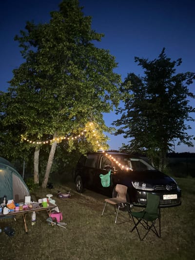 The image depicts a campsite scene at dusk in Завалочыцкі сельскі Савет, Belarus. The environment is a grassy area with several trees. A dark-colored Volkswagen minivan is parked in the center of the frame. A string of warm-toned lights is draped across two trees, illuminating the area. In the foreground, a green tent is partially visible on the left. A foldable table is set up in front of the tent, laden with various items including containers, food items, and cups. Two camping chairs are positioned in front of the car. A small pink stool and a child's scooter are also present on the ground. The sky is a deep blue, indicating twilight. The overall impression is that of a relaxed camping setup.