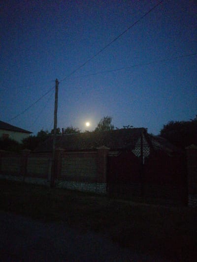 This is a photograph of a rural residential area at dusk or night, with a nearly full moon visible in the dark blue sky. In the foreground, there is a dark, unpaved area. Behind this, a low, brick and stone fence runs horizontally across the image. A wooden utility pole stands to the left of center, with wires extending across the sky. A house with a dark, gabled roof is visible behind the fence, partially obscured by trees. The moon is located in the upper center of the image, to the right of the utility pole and above the roofline. The overall scene suggests a quiet, residential setting in Lanivtsi, Ukraine, characterized by its simple architecture and natural elements under the twilight sky.