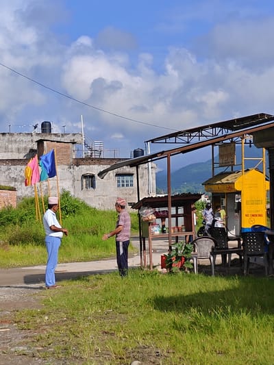This image depicts an outdoor scene in Tharpu, Nepal, featuring two men standing in front of a structure that appears to be a small roadside establishment or a gas station. The man on the left is wearing a light blue short-sleeved shirt, light blue pants, and a patterned cap. He is standing with his hands clasped in front of him, facing the other man. The second man, on the right, is wearing a striped short-sleeved shirt and dark pants, also with a patterned cap. He is facing the first man with his arms slightly outstretched, as if in conversation.
Behind the men, there are three colorful flags (pink, yellow, and blue) mounted on poles. Further back, a building with a concrete facade, several windows, and a water tank on the roof is visible. The environment is characterized by lush green grass and vegetation surrounding the paved area where the men are standing.
To the right of the men, there is a metal-framed structure with a corrugated roof, likely an awning. Underneath this awning, a yellow structure with signage, possibly a fuel pump or service booth, is present. The signage includes text in a script that appears to be Nepali. Chairs are set up in front of this structure, and a motorcycle is parked nearby. The background shows a hilly landscape with trees under a partly cloudy blue sky. The overall impression is that of a rural or semi-urban setting in Nepal, possibly during daylight hours.