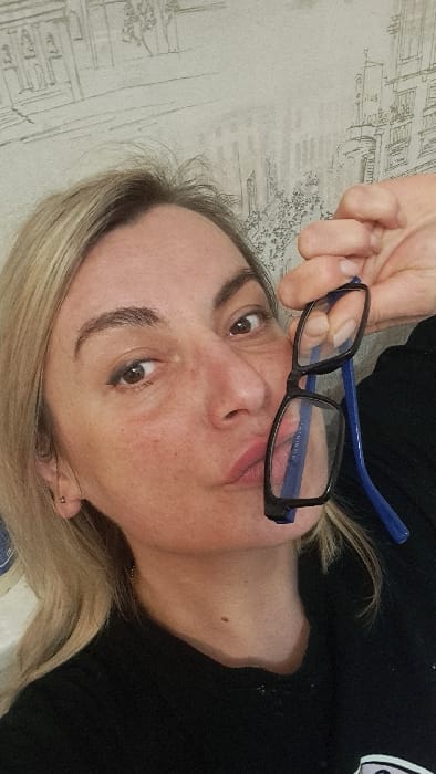 A close-up selfie depicts a blonde female subject indoors in Bel'kovo, Russia. The woman, shown from the chest up, has light-colored hair, dark eyebrows, and eyeliner applied to both eyes. She exhibits some redness on her facial skin. Her lips are pursed forward, and a small, light-colored stud earring is visible in her left earlobe. She wears a dark, possibly black, long-sleeved garment. Her right hand holds a pair of eyeglasses with black frames and blue temples, positioning them in front of her lower face. The background consists of a light-toned wall covered in wallpaper featuring a repeating pattern of sketched monochromatic architectural designs, including buildings and faint tree-like structures.