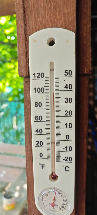 This is a close-up, eye-level, outdoor shot of a white thermometer and hygrometer attached to a wooden post. The thermometer has two scales, one in Fahrenheit on the left and one in Celsius on the right. The Fahrenheit scale ranges from 0 to 120 degrees, and the Celsius scale ranges from -20 to 50 degrees. The red liquid in the thermometer is at approximately 72 degrees Fahrenheit and 22 degrees Celsius. The hygrometer dial is below the thermometer, showing a humidity reading of approximately 45%. The background is out of focus and shows green foliage and a wooden deck or porch. The scene suggests a warm, sunny day. The text visible on the thermometer includes temperature readings and unit labels "°F" and "°C". The hygrometer has text including "HYGROMETER", "%", and numerical values from 0 to 100. There is no specific time of day indicated, but the lighting suggests daytime. The location is identified as Ożarów Mazowiecki, Poland, which aligns with the outdoor setting and the types of trees and foliage visible. No people are present in the image.