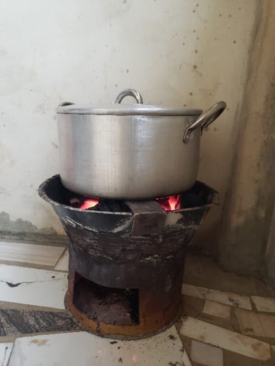 The scene, located in the city of Agunmo, Nigeria, depicts an indoor or semi-sheltered cooking area. A large, lidded, silver-colored aluminum pot with two side handles and a central lid handle is positioned directly on top of a dark, cylindrical metal charcoal stove. Within the stove's upper section, charcoal is actively burning and glowing red. The stove features an open rectangular vent or access point at its base. The floor beneath the stove is covered with white and dark-patterned tiles, some showing signs of wear or discoloration. The background consists of a light-colored, textured wall, exhibiting minor staining and a darker vertical corner element on the right. No individuals are present.
