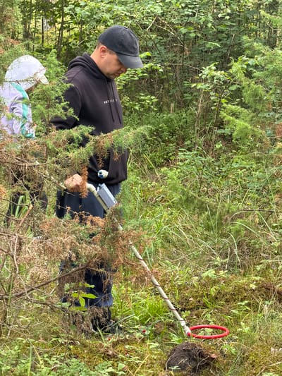 Two individuals are in a forest in Mucenieki, Latvia, engaged in what appears to be a metal detecting activity. A person in the foreground, wearing a black baseball cap and a black hoodie, is holding a metal detector. The device has a grey control box and a circular red coil resting on the ground. To the left, another individual is partially visible, wearing a white cap and a light-colored hoodie with a turquoise accent. They are standing amidst dense vegetation, including trees, bushes, and ground cover such as moss and grass. The environment suggests a natural, wooded setting.