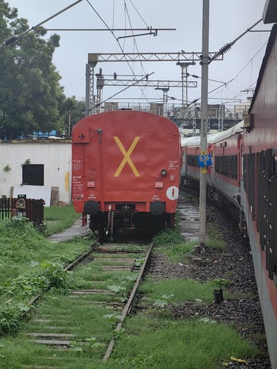 The image depicts a railway track scene in Bhopal, India, under an overcast sky. A red freight car with a large yellow 'X' painted on its side is stationary on the left track. Textual markings on the car indicate it is a RHV 984120, with a tare weight of 37.50 T. A passenger train, predominantly red and white, runs parallel on the right track. Several overhead electrical wires and supporting structures are visible, characteristic of an electrified railway line. The tracks are bordered by lush green grass and some scattered debris. A utility pole with blue signage displaying '836 9' stands between the two trains. The environment appears somewhat unkempt, with vegetation growing around the tracks and equipment. The scene is static, with no visible human activity or movement.