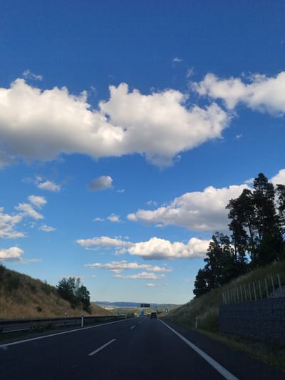 The image captures a view from a highway in Mladotice, Czechia, on a clear day with a bright blue sky filled with scattered white cumulus clouds. The highway stretches into the distance, with two lanes visible. On the left side of the highway, a grassy embankment slopes upward, covered in dry, yellow grass and dotted with sparse trees. A metal guardrail runs along the edge of the embankment. On the right side, another grassy slope rises, featuring a line of tall, dark green coniferous trees at the crest. A gabion wall, filled with rocks and topped with metal fencing, is visible along the side of this slope. A few vehicles are present on the highway, including a dark-colored car further ahead. Overhead, road signs are visible. The overall scene depicts a rural highway environment under a dynamic sky.