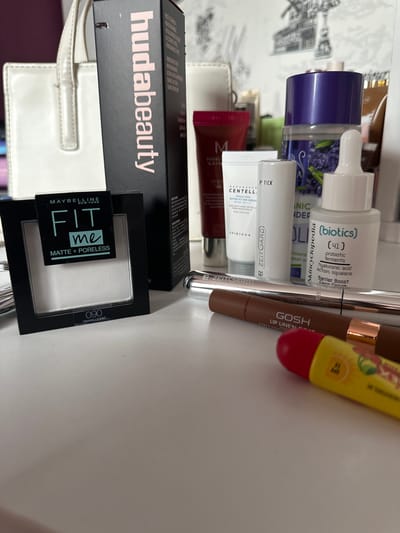 The image shows a collection of cosmetic products arranged on a white surface. The brands represented include Huda Beauty, Maybelline, Skincyclopedia, Skin1004, GOSH and some items without clear labels. The Huda Beauty product is in a black box with pink lettering. A Maybelline Fit Me powder compact is visible in a black case with the shade 090 Translucent. Several skincare items are present, including one labelled "bioticos" from Skincyclopedia, as well as products from Skin1004. A GOSH Lip Liner is also visible. A purse is located on the left of the image.
There is no information available in the image to indicate the city Djerman in Bulgaria or the exact location, scene, or details regarding any people.