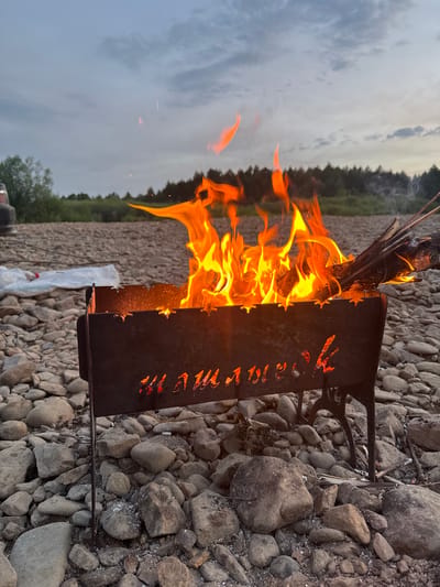 In the city of Pervomayskoye, Russia, an outdoor scene unfolds at dusk. A metal brazier, its sides adorned with cutout Cyrillic letters that read "ШАШЛЫК" (shashlyk), is filled with a vibrant fire. The flames, a mix of oranges and yellows, leap upwards, consuming pieces of wood positioned on the brazier's edge. The brazier rests on a ground covered with an abundance of grey and brown river stones. Behind the brazier, a line of dark green trees forms a dense forest edge against a cloudy, muted sky. To the left of the brazier, a white plastic bag lies on the stones, and the corner of a dark-colored vehicle is visible. The overall environment suggests a relaxed, possibly recreational, outdoor gathering.