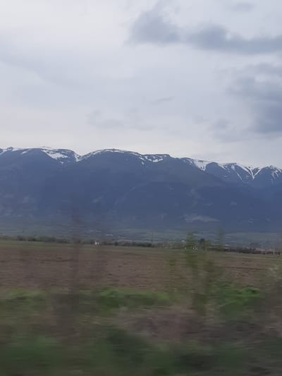 The image depicts a landscape with mountains partially covered in snow on the left, a field in the center, and greenery on the right. The sky is cloudy. Based on the presence of mountains and fields, this could potentially be Marino Pole, Bulgaria, however, there is no confirmation of buildings/infrastructure.