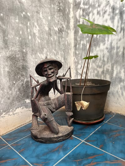 The image depicts an outdoor scene on a tiled floor. In the foreground, a dark wooden carving of a man wearing a conical hat and carrying a yoke on his shoulders is positioned to the left. The carving has a detailed, aged appearance with visible facial features and clothing. To the right of the carving is a dark plastic pot containing a green plant with large leaves and several thin stems. One dried, withered leaf hangs down from a stem inside the pot. The background consists of a rough, textured concrete wall with patches of white paint and grey discoloration, suggesting an aged or weathered surface. The floor is covered in blue tiles with reddish-brown veining. The scene appears to be an outdoor corner, possibly a patio or courtyard in Karangkebagusan, Indonesia.