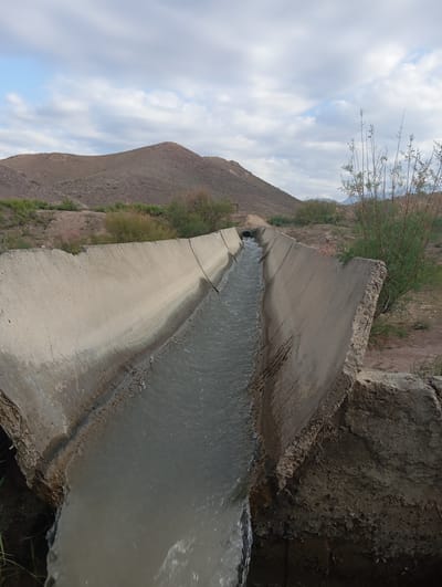 A concrete irrigation canal filled with flowing water cuts through a dry, hilly landscape under a cloudy sky. The canal's edges are uneven and appear to be in disrepair in places. The surrounding environment is arid, with sparse vegetation, including scrubby bushes and dry grasses. In the background, rolling hills dominate the scene, with one larger, more prominent hill to the left. The sky is filled with a mix of light and dark clouds. There are no people visible in the image.