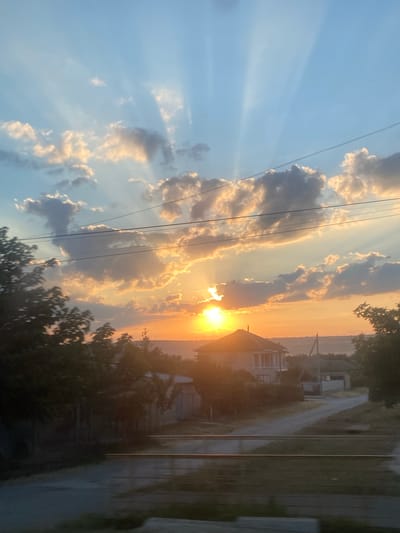 The image depicts a rural sunset scene with crepuscular rays emanating from the sun, which is partially obscured by clouds. In the mid-ground, a two-story house with a dark roof and light-colored walls is visible, situated on a dirt road. Several trees with dark green foliage are scattered throughout the landscape, with some in the foreground appearing blurred due to motion. Overhead, electrical wires are strung across the frame. The background shows a distant, hazy horizon line, suggesting a low-lying landscape. The overall impression is one of a quiet, late afternoon or early evening in a village setting. The location is identified as being in the city of Svetlîi, Moldova.