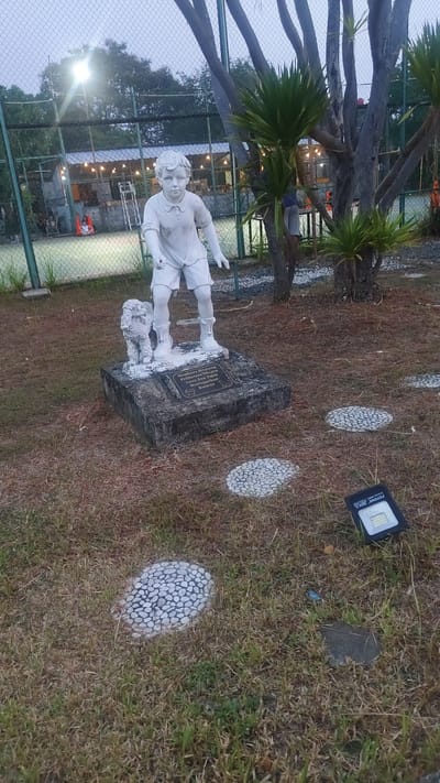 This media file captures a statue of a young boy and his dog in an outdoor setting. The statue, made of white material, stands on a stone pedestal. The boy is depicted in a standing pose, wearing a polo shirt and shorts, with his dog beside him. A plaque is visible on the pedestal, bearing text in Indonesian.

The scene appears to be a park or recreational area, possibly near a tennis court which is visible in the background, enclosed by a green fence. In the distance, there are structures with lights, suggesting a place that is open in the evening or at night. Several circular decorative elements, made of small stones, are embedded in the ground around the statue, forming a path. A solar-powered floodlight is also placed on the ground near the decorative path elements.

The time of day appears to be dusk or early evening, as the sky is overcast and artificial lights are illuminated in the background. The ground is covered in dry grass and fallen leaves, indicating a possibly dry season. The overall mood is serene and contemplative, with the statue serving as a focal point in the tranquil environment.

The visible text on the plaque reads:
"Membawa Binatang
Penasaran Dengan
Anak-Anak Tidak
Meninggalkan
Kotoran"

This translates to: "Bringing Animals Curious About
Children Do Not Leave
Waste".