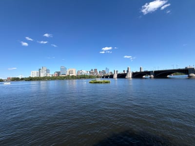 This is a wide, horizontal, eye-level shot of a city skyline across a body of water. The sky is a clear, bright blue with a few wispy white clouds. In the foreground, dark blue water fills the bottom half of the frame, with small ripples on its surface. A small, circular patch of green vegetation, appearing to be an island, floats in the middle of the water.
Beyond the water, a city skyline stretches across the horizon. The buildings vary in height and architectural style, with a mix of modern skyscrapers and older structures. A large, arched bridge with stone towers spans the water to the right of the frame. Several boats are visible on the water, including a white motorboat on the left and a pink object on the water near the bridge. The location is Cambridge, United States of America.