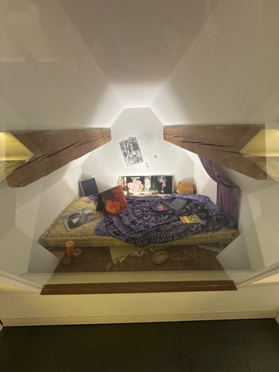 The image displays a staged representation of a bohemian-style room, possibly a recreated historical exhibit. The room features a mattress on the floor with a purple patterned blanket draped over it. Various objects are scattered on and around the mattress, including records, books, a purse, and candles. The space is framed by slanted wooden beams and white walls, suggesting an attic or loft environment. Pictures, including one resembling Janis Joplin, are affixed to the walls. The scene appears to be enclosed in a glass display, indicating it is a preserved or recreated setting. The context of this image has no connection to the city of Bern, Switzerland.