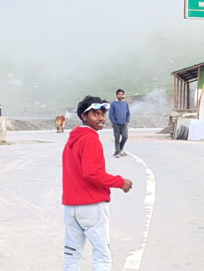 In Marhi, India, two young men are captured in a photographic moment on a paved road. The foreground is dominated by a man wearing a red hooded sweatshirt and light-colored, distressed jeans. He is turned towards the viewer, offering a slight smile, with a cigarette held between his lips and white-rimmed sunglasses perched on his head. His posture suggests a relaxed, informal demeanor.

Further back on the road, another man stands with his hands in the pockets of his dark trousers. He is wearing a blue crew-neck sweater over a white collared shirt, and his gaze is directed away from the camera.

To the left of the men, a light brown cow stands facing away from the camera, its presence indicating a rural or semi-rural setting. In the background, a gentle slope rises, partially obscured by a light, hazy atmosphere. A small, rustic structure with a sloped roof is visible on the right side of the image, with a green sign overhead. Two tires are stacked near the structure. The overall environment suggests a mountainous region, possibly a roadside stop or a village in the Himalayas.