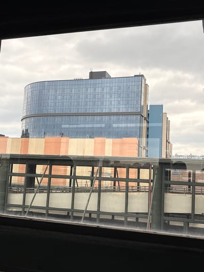 The image shows a view of modern buildings, possibly a complex of offices or residential apartments, seen through a window. The buildings feature extensive glass facades with a grid pattern of small, rectangular windows. The exterior of one building is accented with alternating bands of light beige and salmon-colored panels. A covered walkway or skybridge with a steel frame and glass panels runs in front of part of the buildings, reflecting the architectural details. The sky is overcast and cloudy, suggesting a daytime scene with diffused light. The overall impression is one of urban architecture. There is no discernible text in the image, and no people or specific activities are visible. The orientation of the image is such that the buildings appear to be seen from a slightly elevated position, looking upwards.