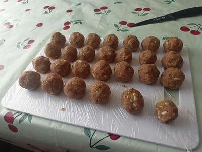 A white cutting board, covered in plastic wrap, holds 25 small, spherical food items, likely meatballs or falafel, arranged in rows. These items are a light brown color and appear to have small pieces of green and white incorporated within them. Some of the spheres contain visible elongated white pieces, possibly pine nuts. The cutting board rests on a table covered with a tablecloth patterned with red cherries and green stems. To the upper right of the cutting board, a black-handled knife with the word "Slice" etched onto its blade is visible on the tablecloth. The lighting suggests it is daytime. The scene appears to be indoors, possibly a kitchen, given the presence of food preparation items.
