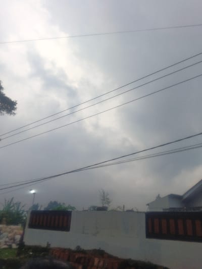 The scene depicts an outdoor environment in Dakshin Hichachhara, India, under a heavily overcast sky composed of dark grey clouds. Multiple utility wires, approximately five to six in number, traverse horizontally across the upper and middle sections of the frame. A dark-foliaged tree is partially visible on the far left of the upper frame. In the mid-ground, a light-colored concrete or plastered wall spans horizontally, featuring two distinct rectangular sections with dark, possibly wooden or tiled, infill. Behind the wall, on the left, a street light with a brightly illuminated white bulb is visible amongst green vegetation, including broad leaves. Further right behind the wall, small trees or shrubs are present. The upper portion of a building with a light-colored, possibly grey, sloped roof is visible on the far right, partially obscured by the wall and vegetation. The lower foreground is dark and indistinct, containing reddish-brown objects resembling bricks or rubble. No individuals are present in the image. The lighting is dim and diffused.