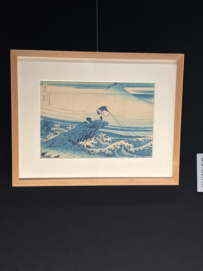 The image displays a framed Japanese woodblock print depicting a scene with Mount Fuji in the background. In the foreground, a figure, likely a fisherman, is shown standing on a rocky outcrop amidst turbulent waves, casting a net into the water. The print is matted and enclosed in a light-colored wooden frame. Below and to the right of the frame, a small informational label is visible with Japanese text and some English translation, indicating it is an exhibit, possibly at a museum or gallery in the city of Sumida, Japan.