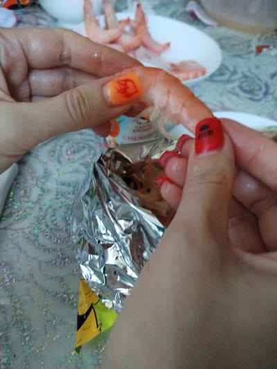 A close-up shot shows two hands preparing shrimp. The hand on the left is holding a shrimp and a small, foil-wrapped packet. The fingers of this hand are adorned with bright orange nail polish with a red flame design. The hand on the right is holding the foil packet open, revealing shrimp inside. The fingers of this hand have bright red nail polish. In the background, a white plate holds several cooked shrimp. The surface beneath the hands has a sparkly, patterned texture. The image appears to have been taken indoors.