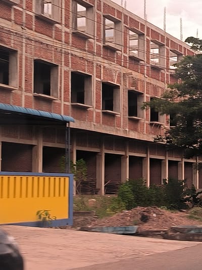 The image captures a multi-story building, likely under construction or recently completed but unoccupied, situated in Lampeuneurutgampong, Indonesia. The building's facade features a grid of light gray concrete columns and beams, with sections filled by reddish-brown brickwork. The upper floors display numerous rectangular window openings that are currently unglazed. Rebar extends upwards from the top level, indicating unfinished construction. Several power lines run horizontally across the building's exterior.

The ground floor consists of multiple arched openings, which appear to be either closed off or deeply recessed and darkened. A blue corrugated metal awning extends over the ground-level facade on the left side of the building. In the immediate foreground on the left, a distinct yellow gate or fence with vertical slats and a blue frame is prominent. Further to the right in the foreground, a large pile of dirt and debris, possibly construction waste, is visible, with sparse vegetation growing around it. Mature trees with dense green foliage partially obscure the right side of the building.

The scene is illuminated by diffused daylight, suggesting an overcast sky or a time of day with indirect sunlight. No people are visible, nor is any discernible text present on the building or surrounding structures. A small portion of a dark vehicle is visible in the bottom-left corner.