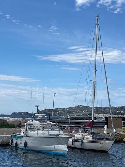 The image shows two boats docked in a harbor. The boat on the left is a white fishing vessel with a blue stripe along the waterline. It has several antennas and a small cabin. The boat on the right is a sailboat with a tall mast and a red sail cover. The name "Mijau" is visible on the hull of the sailboat. In the background, there is a rocky coastline with buildings and vegetation. The sky is blue with scattered clouds. The scene appears to be in Le Lavandou, France, a coastal town known for its beaches and harbor.