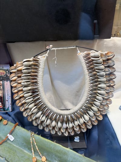 This image shows a display of necklaces in what appears to be a shop window. The primary object of focus is a large, ornate necklace made of numerous cowrie shells, arranged in multiple rows around a thick, woven rope. The shells are a mix of cream, brown, and white, with varying patterns. The necklace is suspended from a dark metal frame. To the left, partially visible, is a decorative item with images of buildings and figures, possibly a piece of artwork or a decorative box. In the foreground, resting on a broad, green, spiky leaf, are two other necklaces. One is made of small, brown, spherical beads and has a price tag attached. The other is a delicate strand of small, multi-colored beads and charms. The background consists of draped fabric in shades of beige and dark blue, and a black, reflective surface which may be part of the shop's display or a window. The overall impression is a retail setting, likely a souvenir shop or a boutique selling handcrafted jewelry. The specific location within Würzburg, Germany, cannot be determined from the visual information provided.